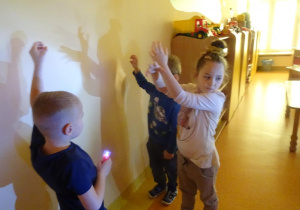 Troje dzieci tworzy cienie ze swoich dłoni na ścianie.
